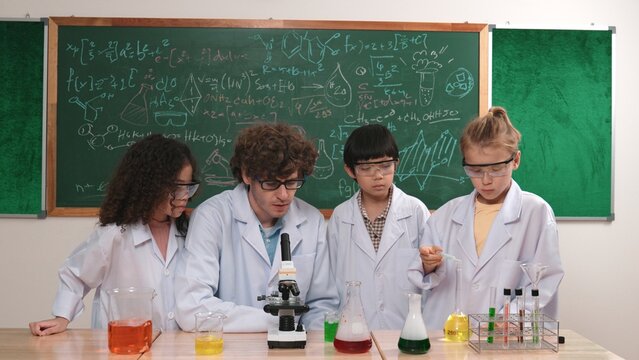 Smart caucasian teacher looking under microscope while diverse children doing experiment at STEM science lesson. Professional happy scientist inspect biochemical example by using microscope. Pedagogy.