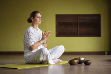 Fototapeta premium Woman practicing Kundalini yoga, sitting in comfortable position on mat and doing lotus mudra from loneliness, practicing in white clothes in room with meditation bowls placed nearby