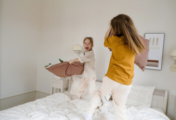 Two sisters are having a playful pillow fight on a bed, laughing and having a great time.