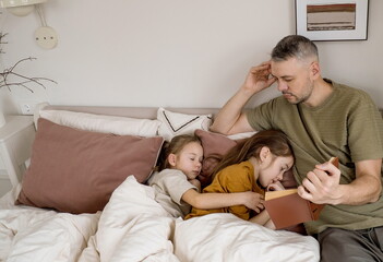 Fototapeta premium A father reads a bedtime story to his two daughters who are already asleep in bed.