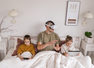 Obraz premium A father sits on a bed, wearing a VR headset, and interacting with the virtual world. His two daughters sit beside him, one using a phone, the other using a tablet.