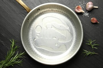 Frying pan with cooking oil, rosemary, garlic and salt on dark textured table, flat lay