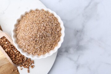 Buckwheat bran in bowl and grains on white marble table, top view. Space for text