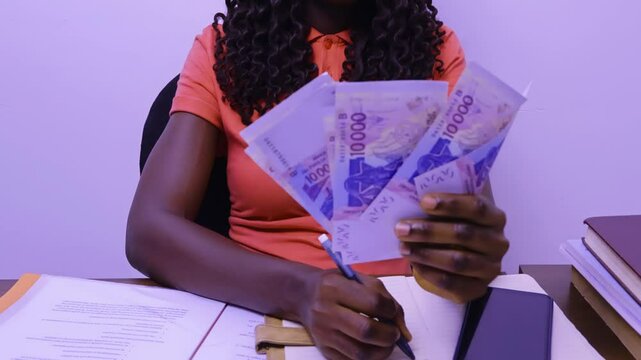  Young black african female lady woman wearing orange shirt top sitting at office desk holding counting West African CFA franc notes