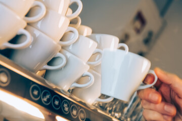 Barista is seen grasping a white coffee cup from a tower of stacked cups in a lively café. Customers gather for their morning drinks, creating an inviting atmosphere.