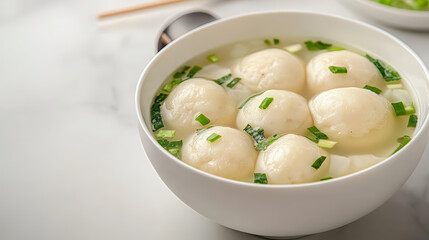 A bowl of soup with dumplings, garnished with chopped green onions, placed on a white table with chopsticks and a spoon nearby.