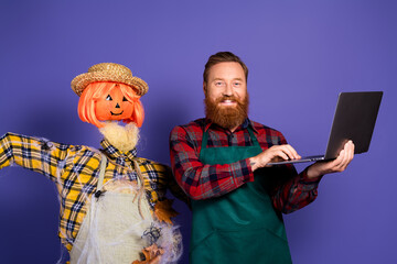 Photo of cheerful man use device standing near scarecrow doll guarding field isolated on purple color background