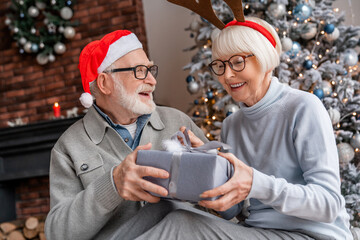 Happy senior couple in Santa hats exchanging Christmas gifts at home - Powered by Adobe