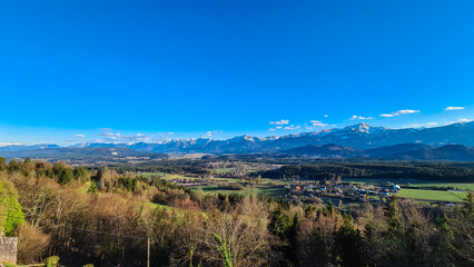 Panoramic view of snow-capped mountain range Karawanks in Carinthia, Austria, Europe. Alpine terrain of Austrian Alps surrounded by pine tree forest. Dramatic sky over winter wonderland landscape