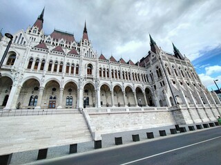 A dramatic wide-angle view of the Hungary Parliament building's neo-Gothic front façade. The image captures the intricate details of pointed arches, elaborate spires.