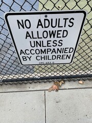 This photo captures a sign posted at a children's playground that reads, "No Adults Allowed Unless Accompanied by Children." The sign is mounted on a chain-link fence.