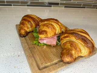 A delicious croissant sandwich filled with ham and lettuce, presented on a wooden cutting board in a modern kitchen setting. A medium-angle shot with a neutral background.