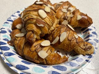 Croissants topped with sliced almonds and drizzled with chocolate, displayed on a patterned paper plate. A perfect treat for dessert or breakfast.