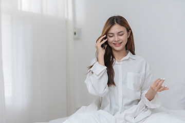 Young asian woman is relaxing in bed on a weekend morning, having a pleasant phone call
