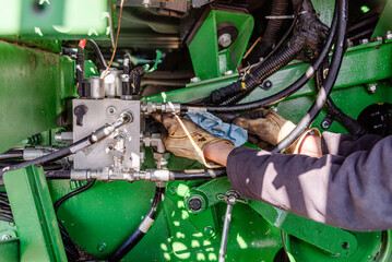 Farmer Mechanic Repairing Hydraulic Hose Under Combine Undercarriage, Wearing Yellow Gloves, Using Wrenches, Propped by Dual Tires, Agricultural Equipment Maintenance, Combine Harvester Repair