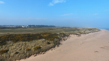 Aerial view of coastal landscape. Sandy beach, colourful houses in distance and golf course visible on the hillside