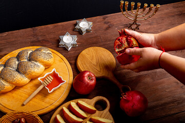 Female hands breaking a pomegranate on the table in Rosh Hashanah next to an apple challah honey...