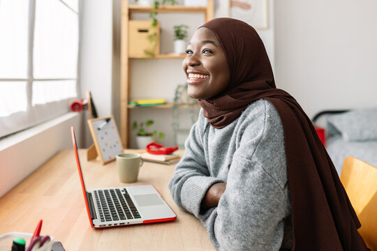 Smiling young dreamy african muslim woman with laptop sitting at desk in her room. Happy black female student in muslim headscarf relaxing at home. Youth and technology concept.