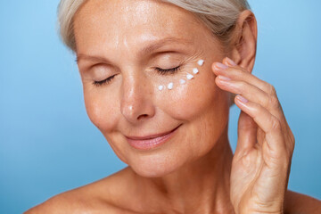 Smiling mature woman with closed eyes applying moisturiser under her eyes, showing a relaxed skincare routine against a blue background...