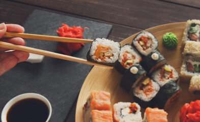 Japanese food restaurant, sushi maki roll plate, platter set. Hand take roll. Set for two with chopsticks, ginger, soy, wasabi. Sushi at rustic wood background and black stone. POV at black. Closeup.