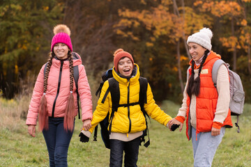 family walking in autumn forest with backpacks, boy in yellow jacket, girl in pink jacket, mother in orange sleeveless jacket, family in hat with pompoms, gen z