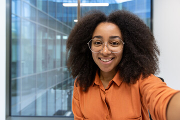 Smiling woman with curly hair taking a selfie while wearing glasses in brightly lit modern office building