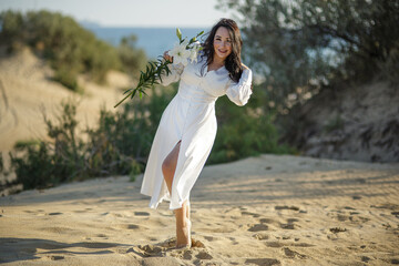 Beautiful brunette girl posing on a sandy beach in a sexy white dress with a flower in her hands.