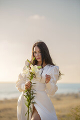 Beautiful brunette girl posing on a sandy beach in a sexy white dress with a flower in her hands.