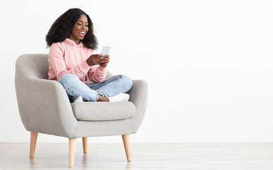 Cheerful african american young lady sitting in arm chair at home, using nice mobile application on...