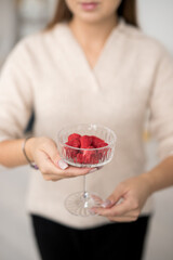 Delightful raspberries presented in a crystal glass by a woman in a cozy, neutral-toned setting on a bright afternoon