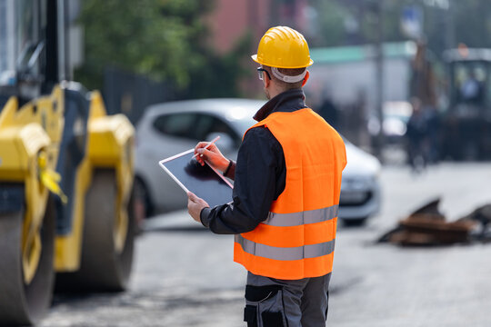 Construction worker in a safety vest using a tablet on a busy street during the day near heavy machinery and parked vehicles - Powered by Adobe