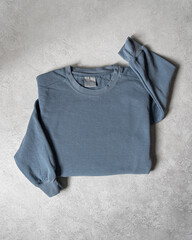 Blank Light Blue Crewneck Sweatshirt Folded on Concrete Background, Stylized Sleeves, Natural Lighting with Bold Shadows, Simple Product Shot with Blank Tag, Casual Apparel Photography Mockup Template