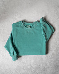 Blank Light Green Crewneck Sweatshirt Folded Concrete Background, Stylized Sleeves, Natural Lighting with Bold Shadows, Simple Product Shot with Blank Tag, Casual Apparel Photography Mockup Template
