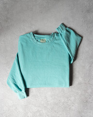 Blank Teal Blue Crewneck Sweatshirt Folded on Concrete Background, Stylized Sleeves, Natural Lighting with Bold Shadows, Simple Product Shot with Blank Tag, Casual Apparel Photography Mockup Template