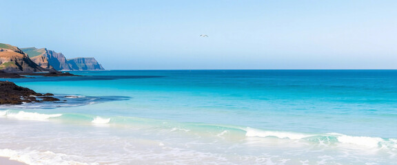 Obraz premium Serene ocean scene with clear blue waters gently lapping a sandy beach, with rugged cliffs in the distance. Ideal for themes of tranquility, natural beauty, relaxation, and coastal landscapes.