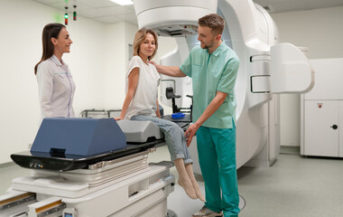 Patient sits on linear accelerator table, preparing for radiotherapy, assisted by two healthcare...