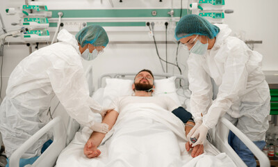 A male patient lies in a hospital bed with an oxygen mask, while two healthcare workers in full...