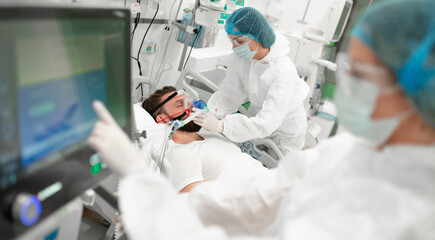 Healthcare worker in full protective gear adjusts breathing mask on male patient in hospital bed,...