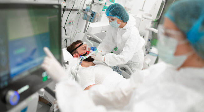 Healthcare worker in full protective gear adjusts breathing mask on male patient in hospital bed, surrounded by medical equipment, highlighting critical care, respiratory support, patient treatment