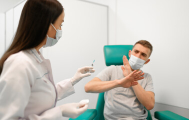 Masked healthcare professional prepares to administer vaccine, while nervous male patient raises his hands in apprehension. The image captures vaccine anxiety and healthcare in a clinical setting