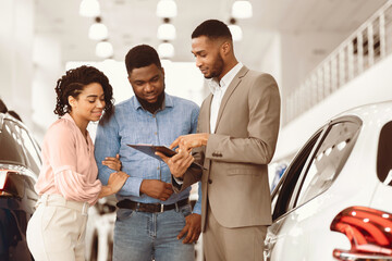 Car Sales. Afro Spouses Buying Auto Consulting With Dealer Guy Standing In Luxury Dealership Store.