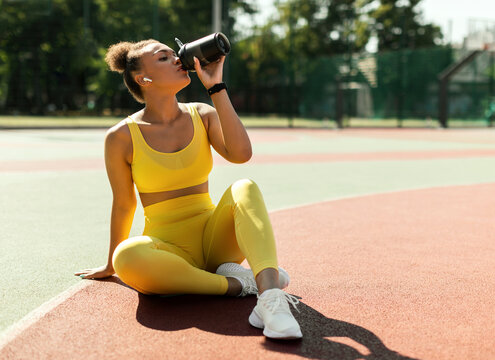 Energy, Vitality, Wellbeing Concept. Fit African American woman in wireless earbuds sitting on basketball field ground, holding shaker, drinking water or protein, taking break after outdoors exercise