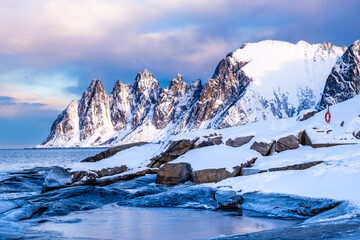 Tungeneset or Devils Jaw mountain on Senja island, Norway in winter. Winter landscape with snowy mountain peaks, fjord waters and ice coast in northern Norway. Scenic view with dramatic sky
