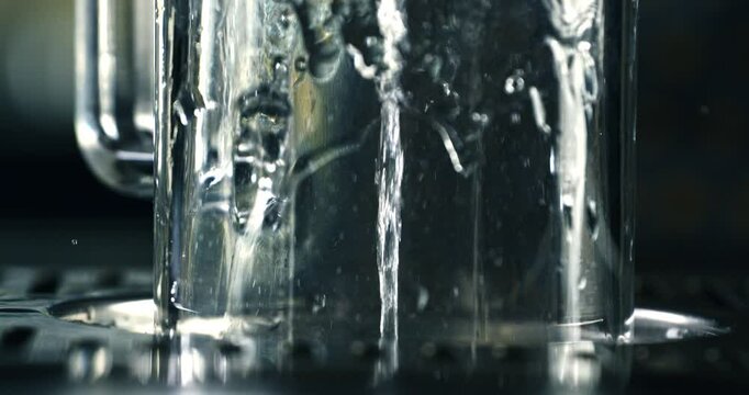 Super slow motion macro of barman or bartender washing transparent beer mug with automatic glass rinser with water spray and splashing drops in bar at 1000 fps.