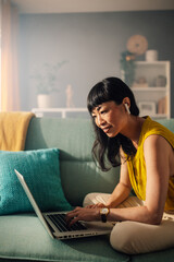 Asian woman on a sofa working on a laptop, surrounded by cushions.