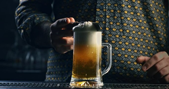 Super slow motion macro of barman or bartender removing froth from foamy golden icy fresh cold beer poured in transparent mug before serving it to customer at bar pub at 1000 fps.