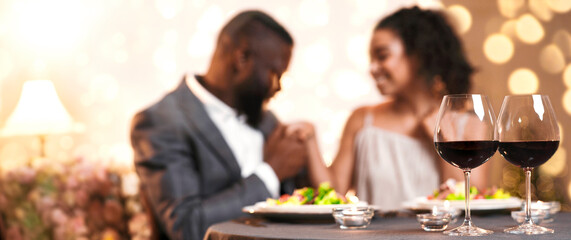 Valentine dinner. Glasses of wine over black man in suit courting pretty young woman, eating meal in restaurant