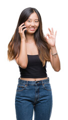 Young asian woman speaking on the phone over isolated background doing ok sign with fingers, excellent symbol