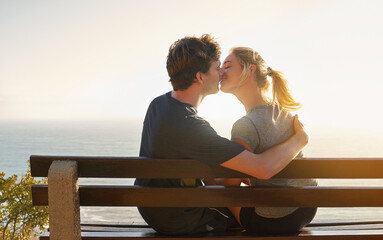 Fototapeta premium Love, kiss and couple at sea at sunset for date, hug or bonding together on bench on holiday. Happy, man and woman at ocean for romantic relationship, support and connection with partner on vacation