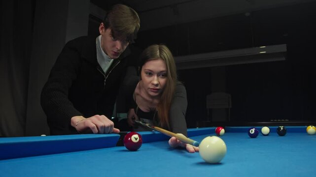 Young man teaches his beloved to play billiards. He talks in detail about rules of the American pool and helps her hold cue. In process of learning, he gently hugs her.